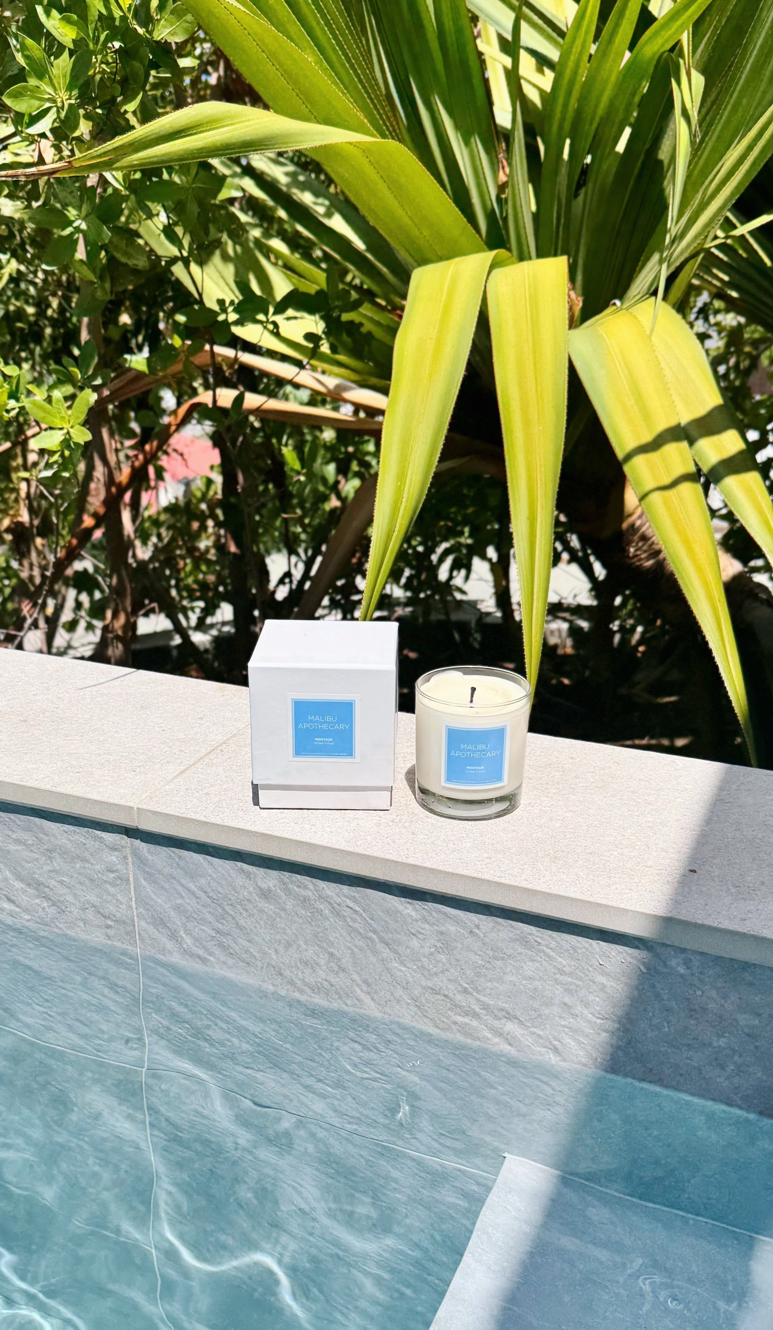 Two clean candles with blue labels on a ledge by a pool with greenery in the background in Saint Barths in the Mykonos scent