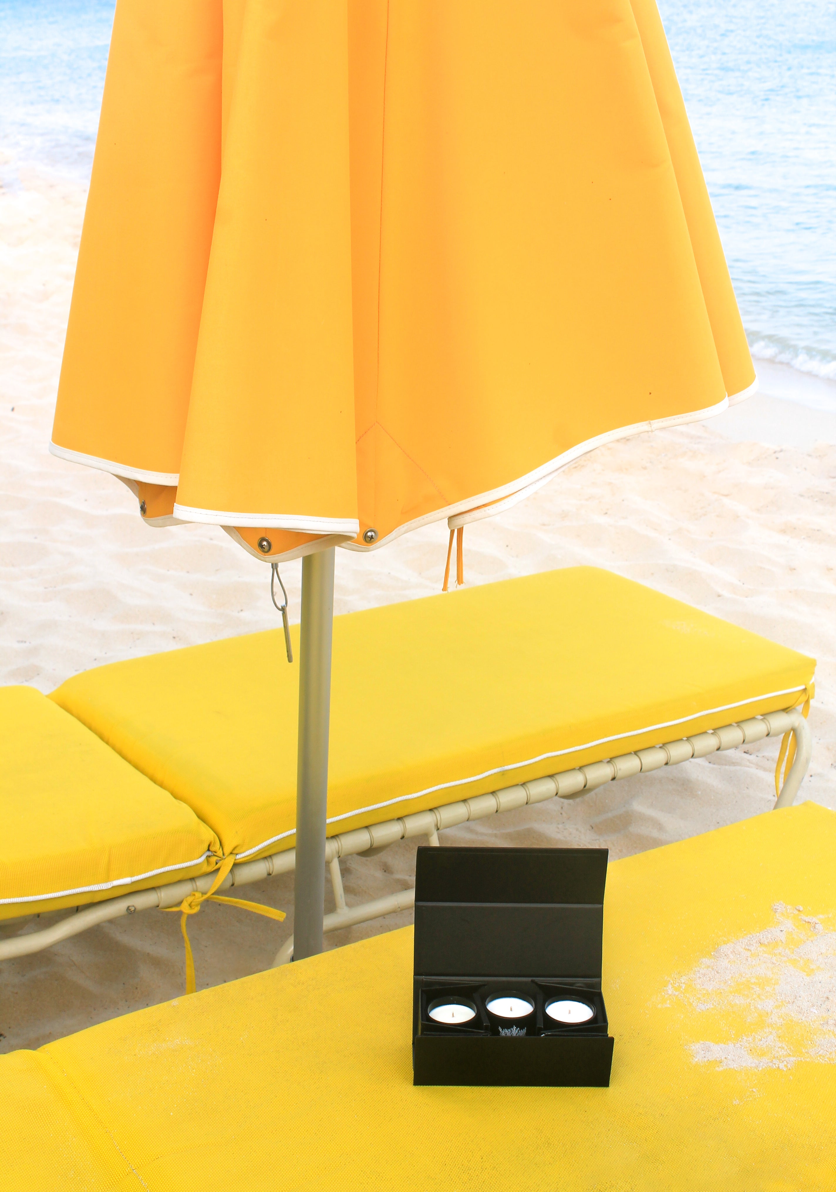 Open black gift box with three mini candles resting on yellow beach lounge chair under umbrella near ocean, hand-poured with all natural coconut and soy wax for luxury gifting and scent marketing