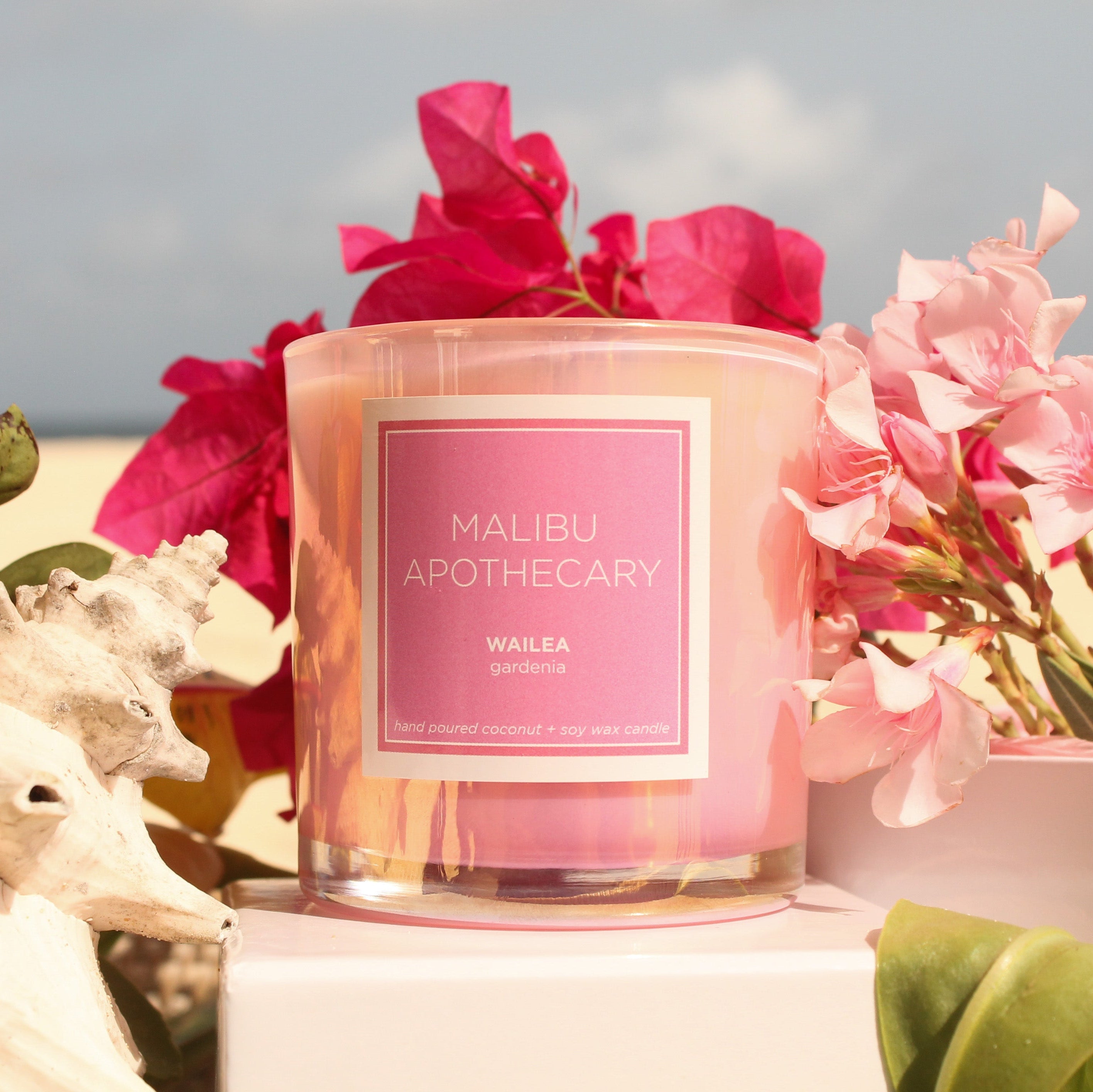 Pink candle with 'Malibu Apothecary' label surrounded by flowers and seashells on a beach in Anguilla