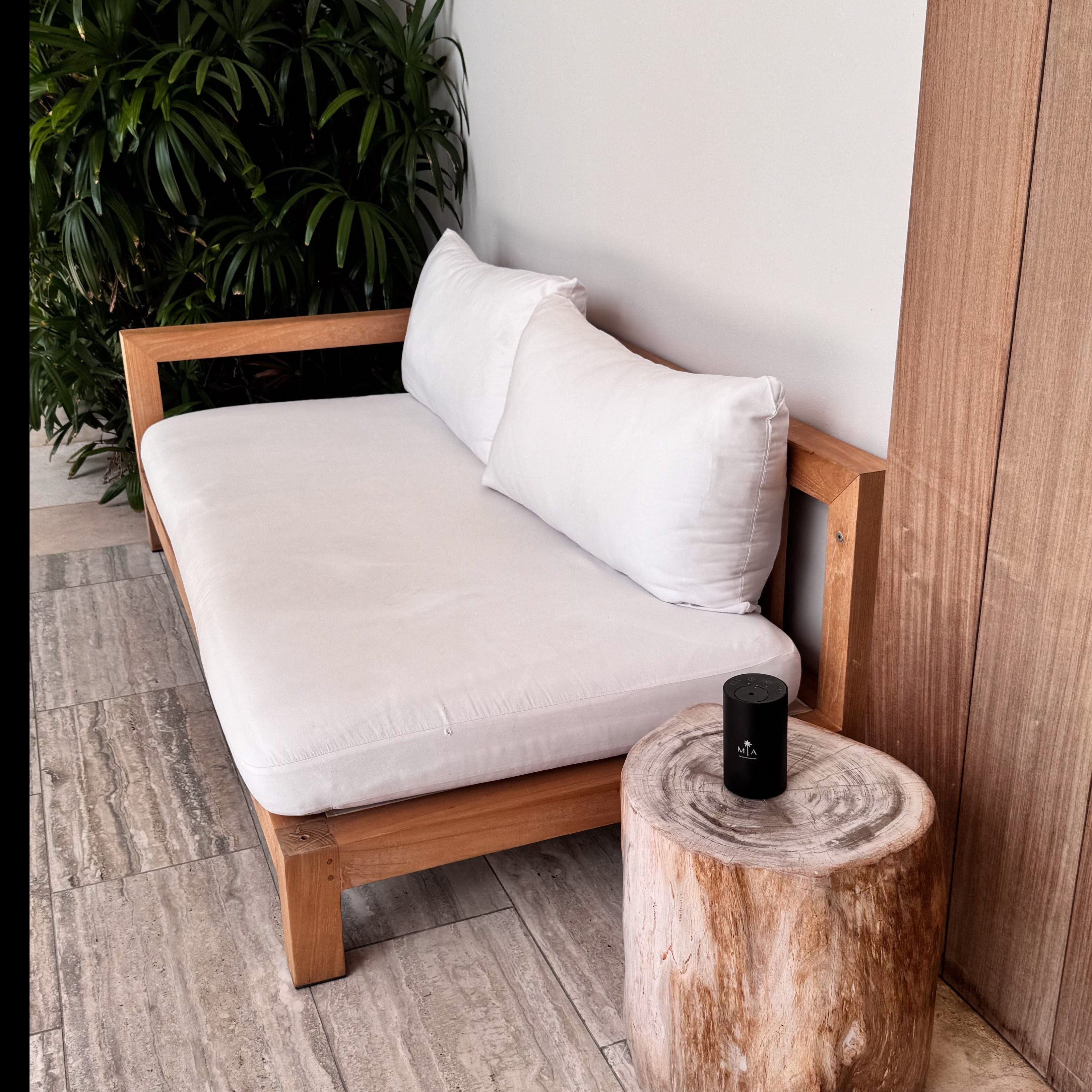 ScentSolution Mini portable scent diffuser in matte black cylinder resting on a rustic wooden stump side table beside a modern outdoor sofa with white cushions and lush potted palm plants; perfect for home or patio aromatherapy.
