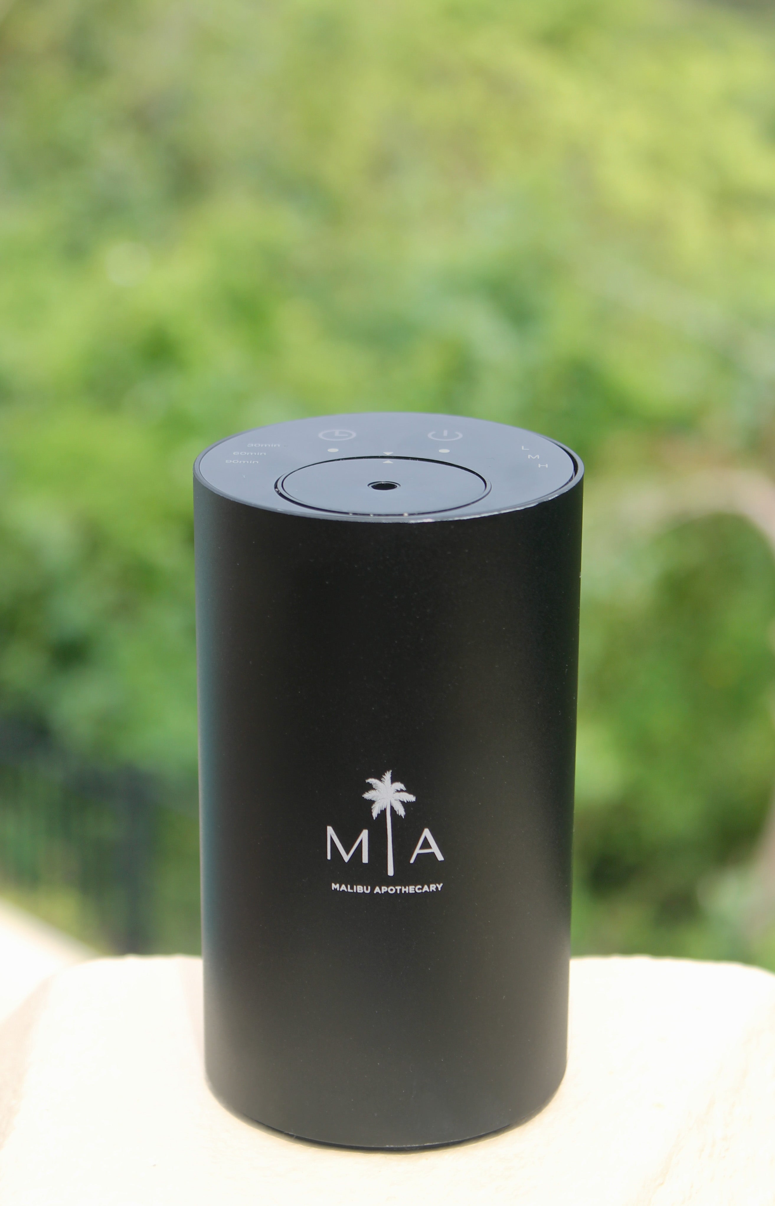 ScentSolution Mini black cylindrical portable scent diffuser with white MIA logo, placed on a wooden surface outdoors against a blurred green foliage background; uses advanced nebulization technology for pure essential oils.