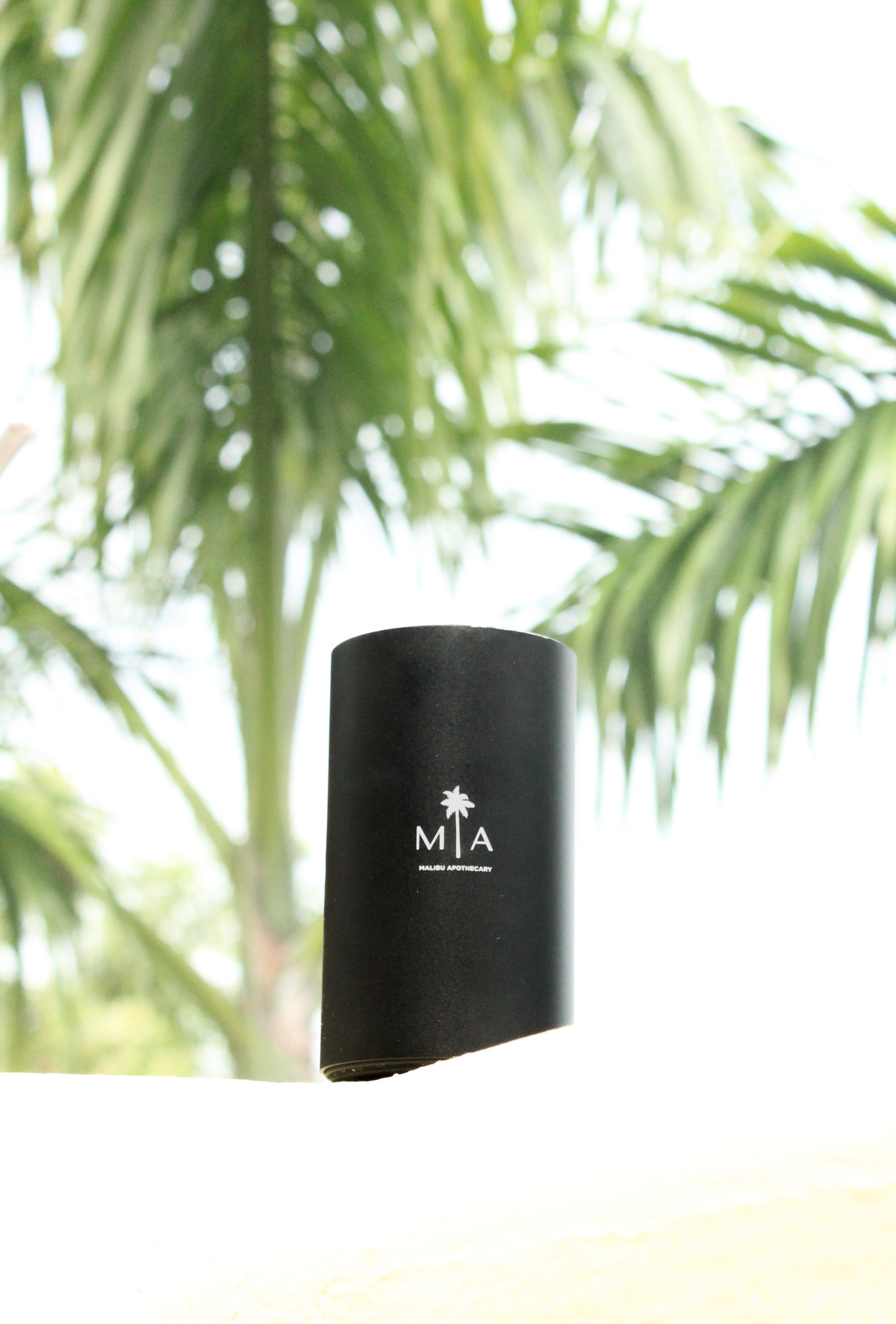 ScentSolution Mini portable black scent diffuser with white MIA logo on a white ledge against blurred tropical palm leaves, capturing an airy, beach-inspired ambiance; ideal for diffusing pure aroma oils.