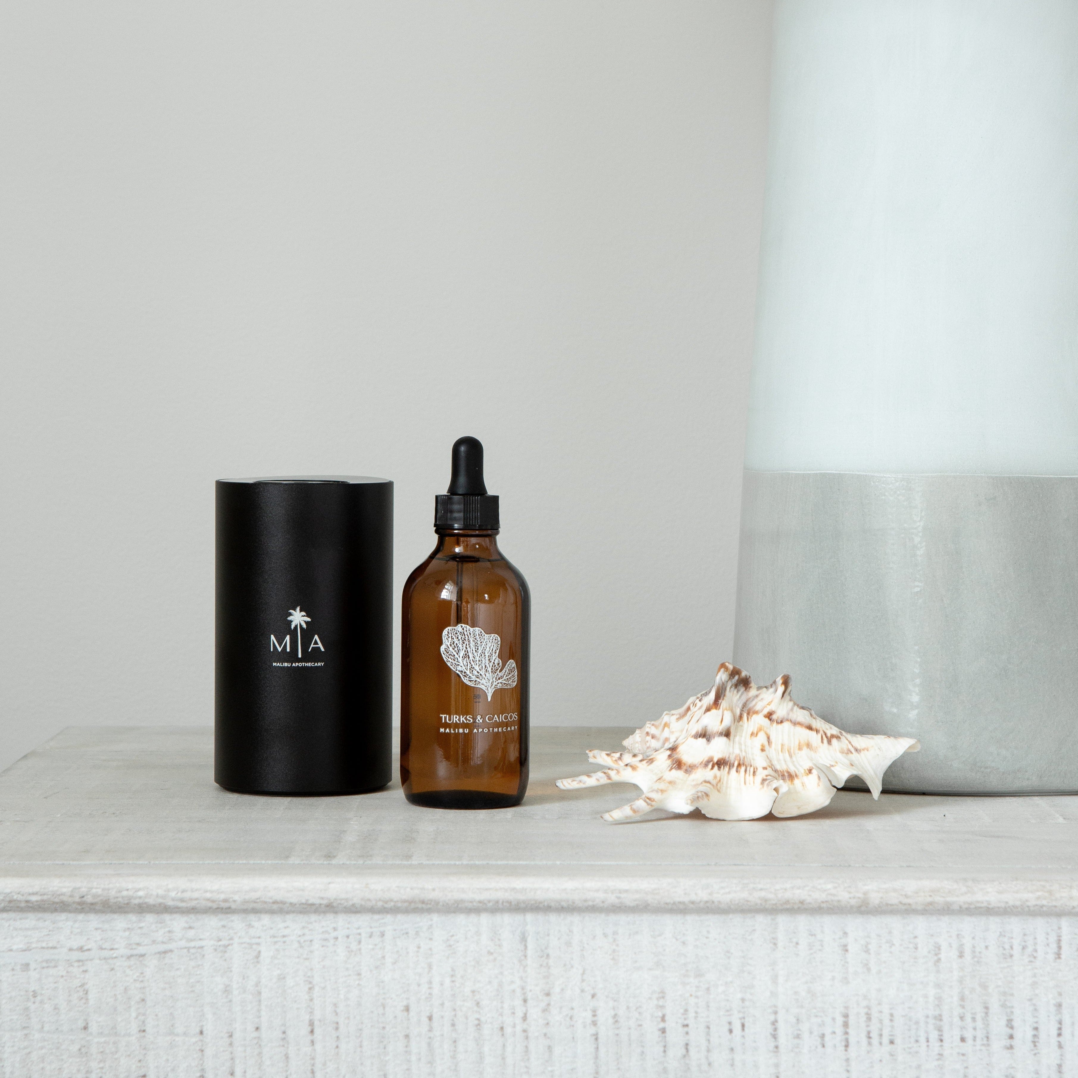 ScentSolution Mini diffuser alongside Bora Bora pure aroma oil dropper bottle and a large conch shell on a gray stone surface with neutral backdrop; designed for use with Malibu Apothecary's scent diffuser system.