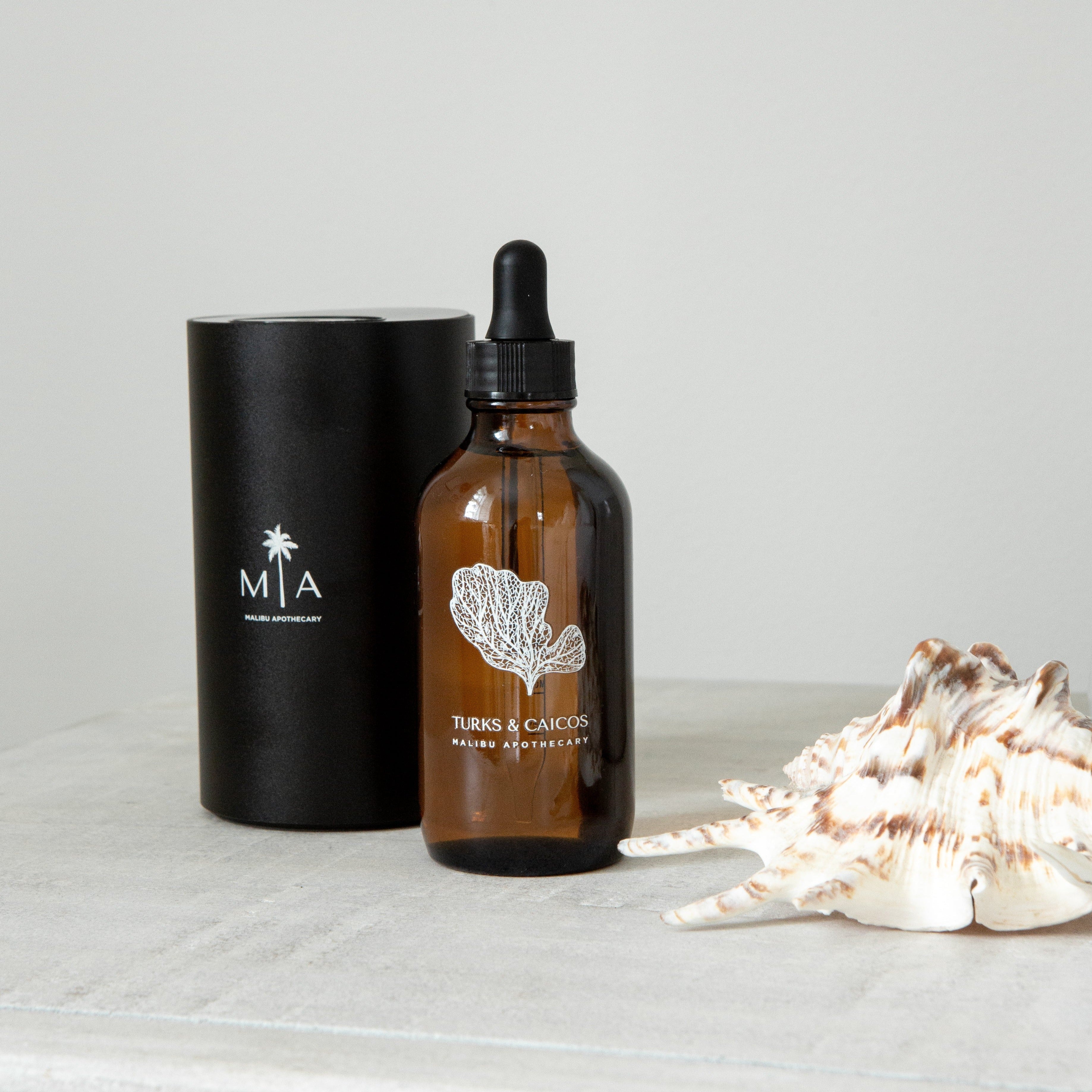 Turks & Caicos pure aroma oil in amber glass dropper bottle with coral illustration, displayed next to black MIA scent diffuser and a large seashell on a concrete surface; designed for use with the ScentSolution Mini nebulizing diffuser.