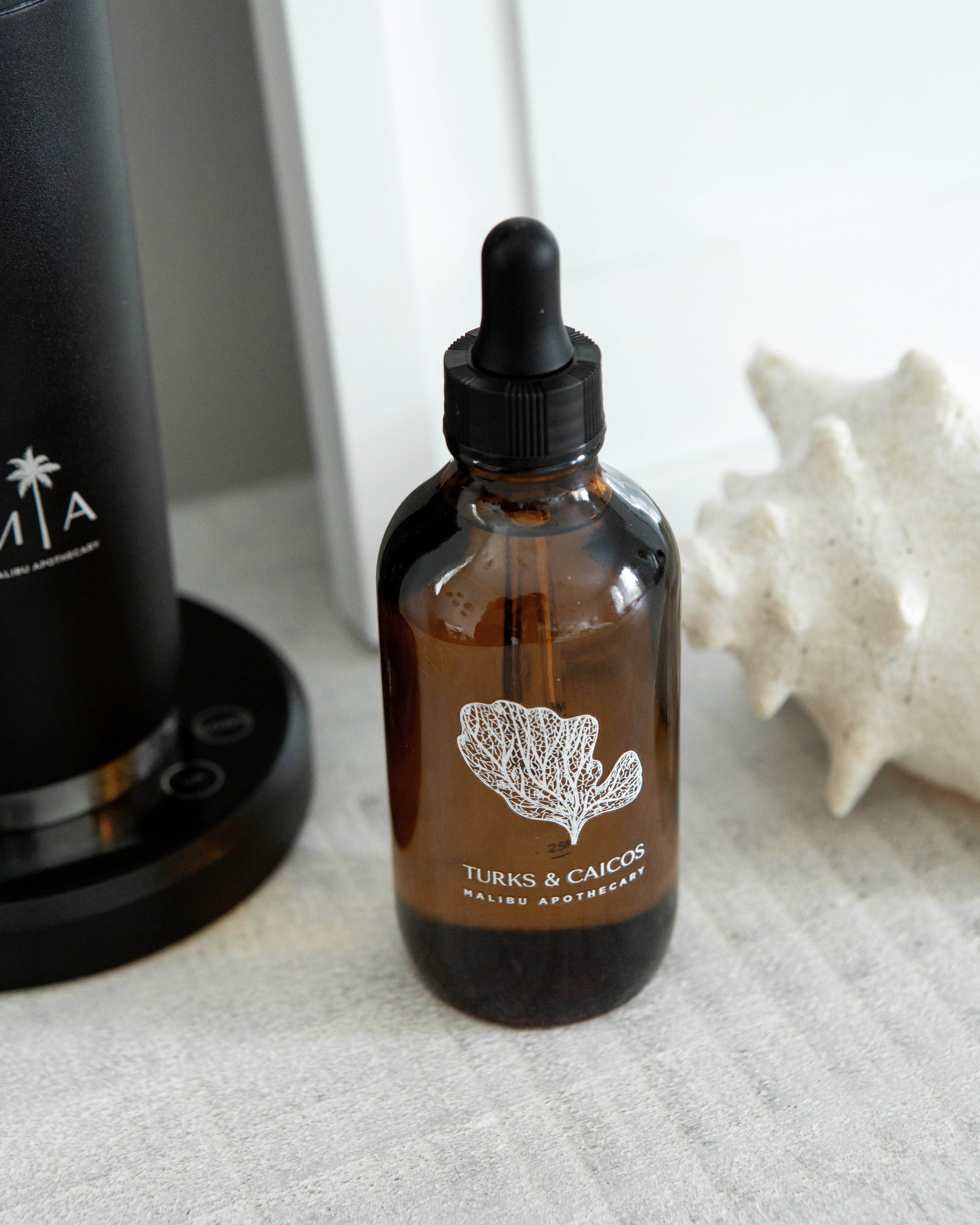 Turks & Caicos pure aroma oil bottle with coral illustration beside scent diffuser base and seashell on neutral surface; perfect for luxury home scenting and spa aromatherapy