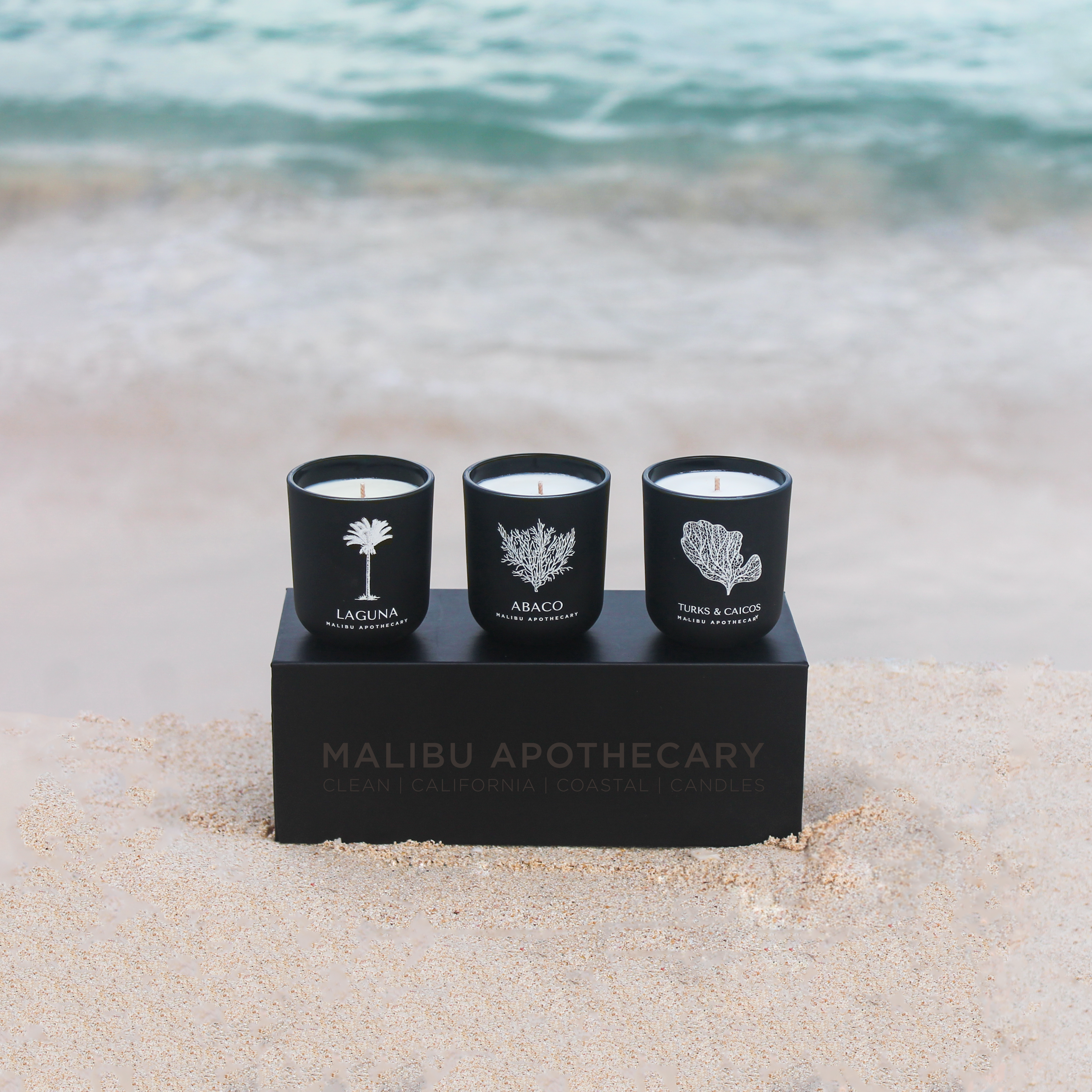 Luxury votive discovery set of three matte black candles on a black box placed on sandy beach with turquoise waves, hand-poured with all natural coconut and soy wax for clean home fragrance