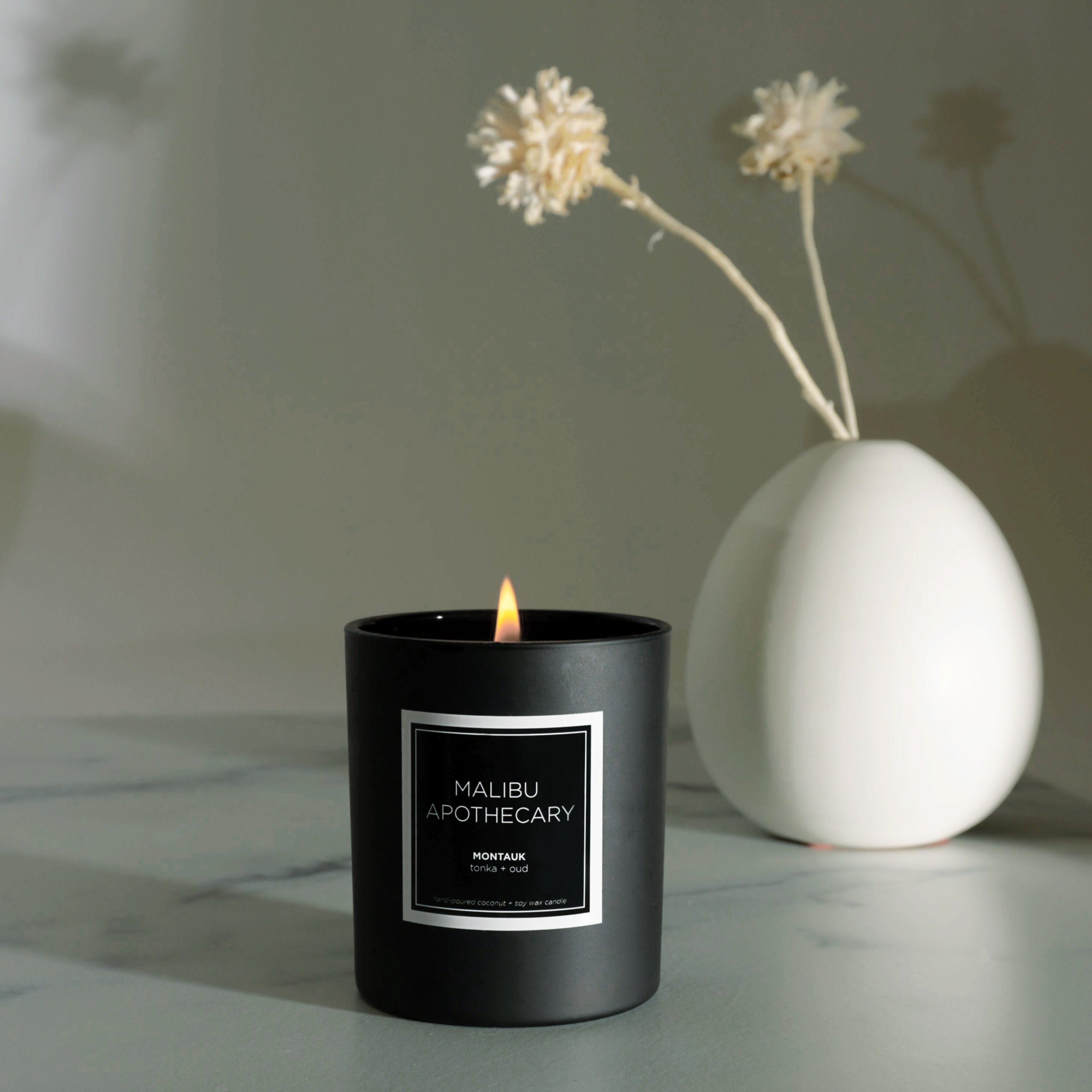 Matte black Malibu Apothecary candle burning on a marble surface beside a white ceramic vase with dried flowers, handcrafted with all natural coconut and soy wax