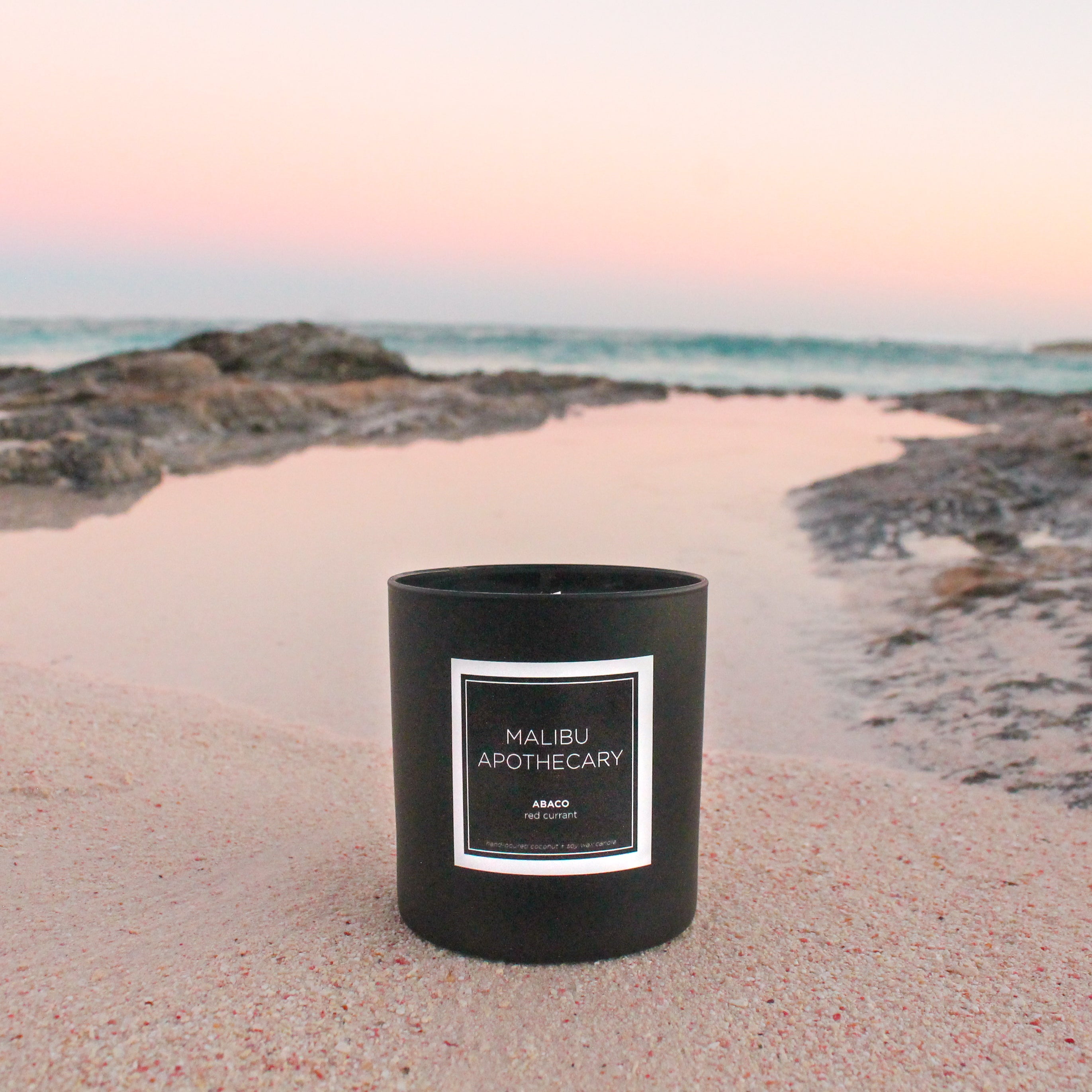 Matte black Malibu Apothecary candle placed on a pink sand beach in Eleuthera, Bahamas with turquoise surf and rocky shoreline, made with all natural coconut and soy wax and featuring red currant fragrance