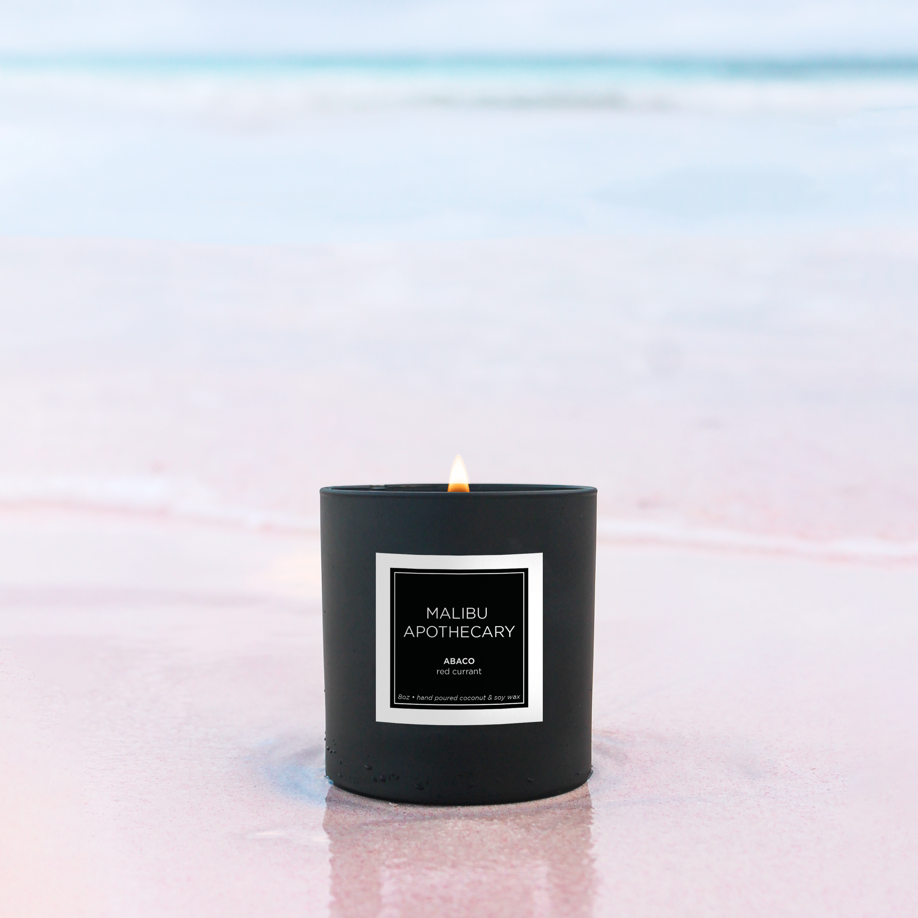 Matte black Malibu Apothecary candle burning on pink sand beach with blurred turquoise ocean in background, made with all natural coconut and soy wax