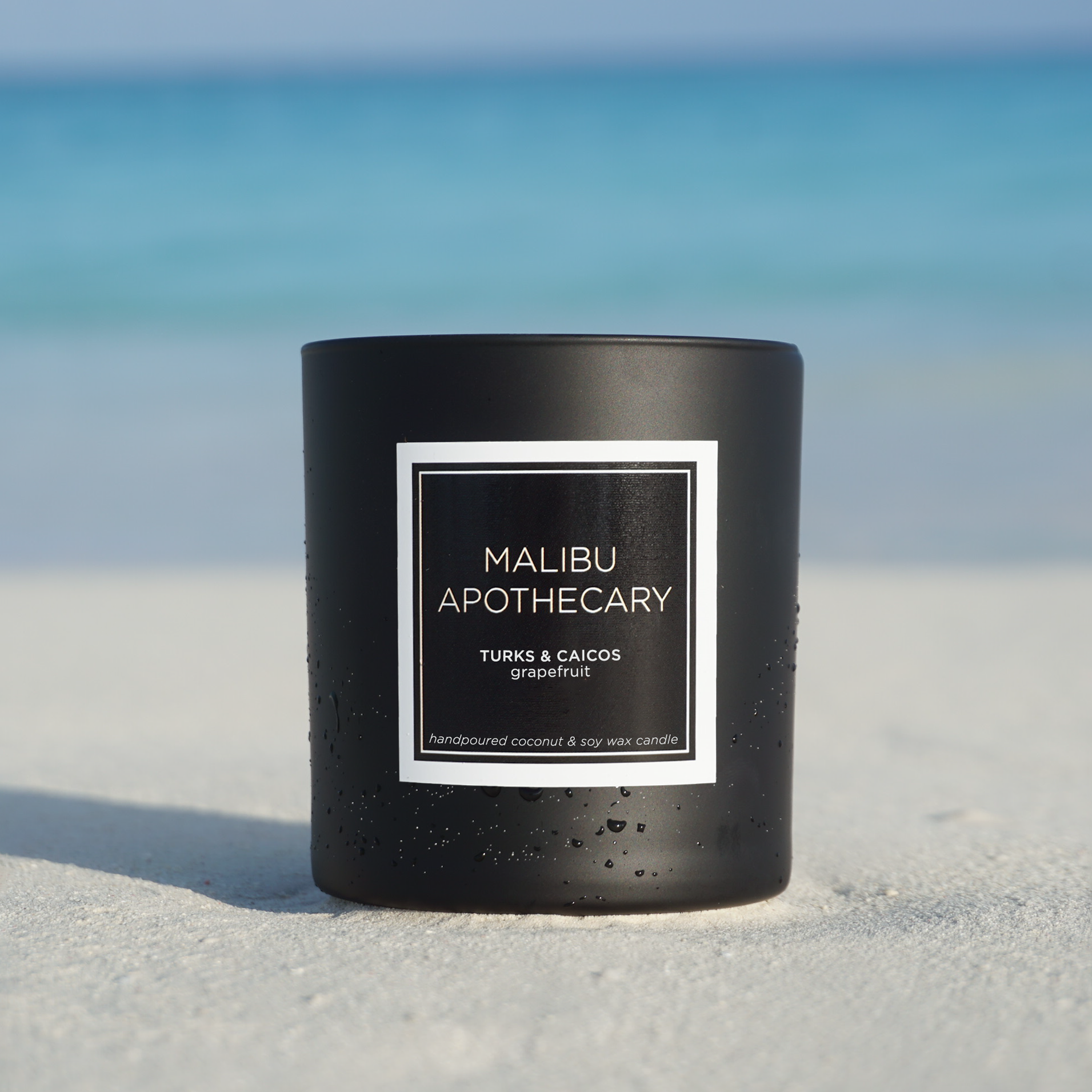 Matte black Malibu Apothecary candle labeled Turks & Caicos grapefruit resting on white sand beach with turquoise water and blue sky, made with all natural coconut and soy wax