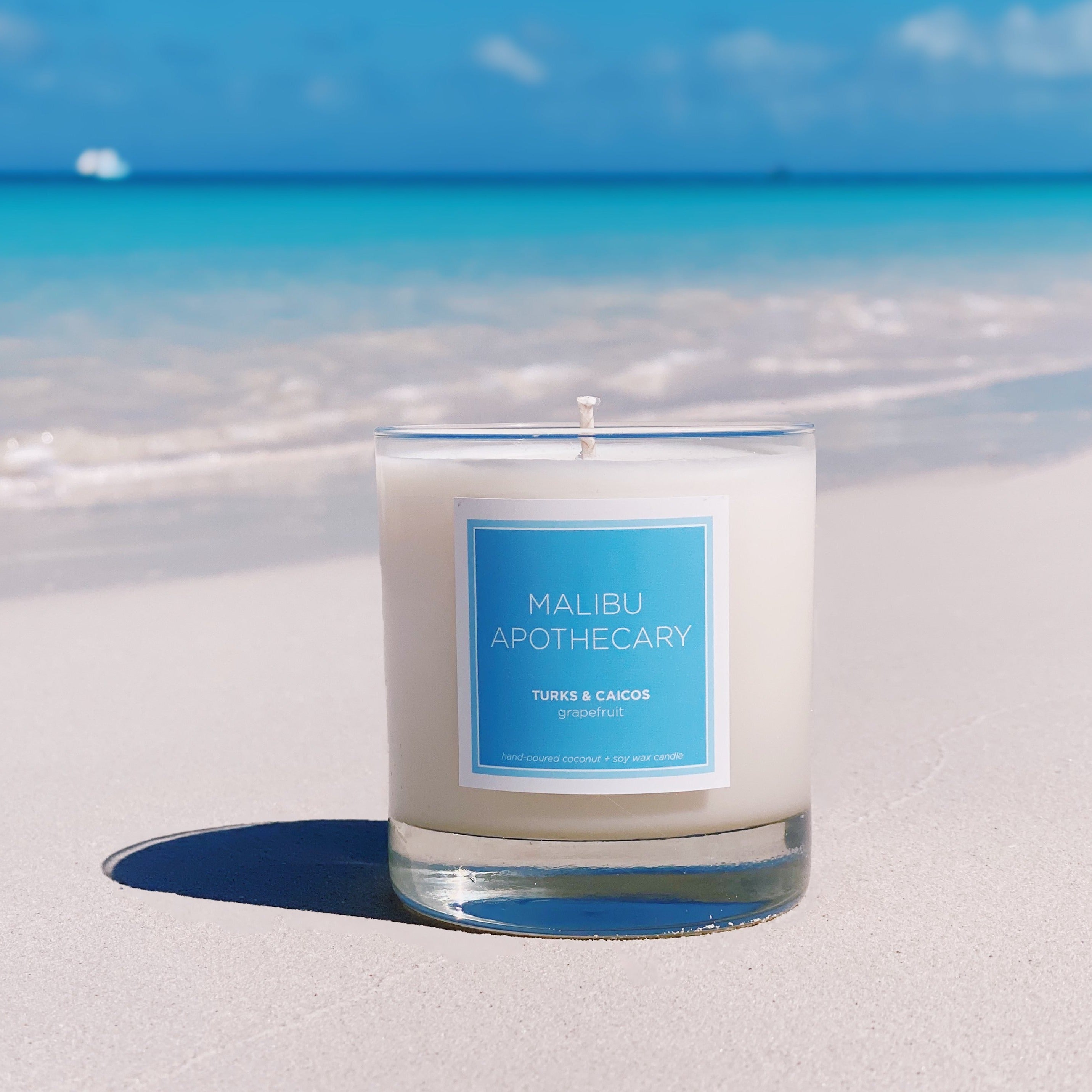 Malibu Apothecary Candle in Clear Gloss and Blue label on the sand in front of the waves in Turks & Caicos