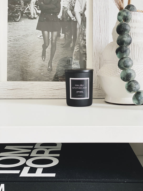 Mini matte black travel candle next to mala beads, a vase, and flowers on a coffee table and Tom Ford book