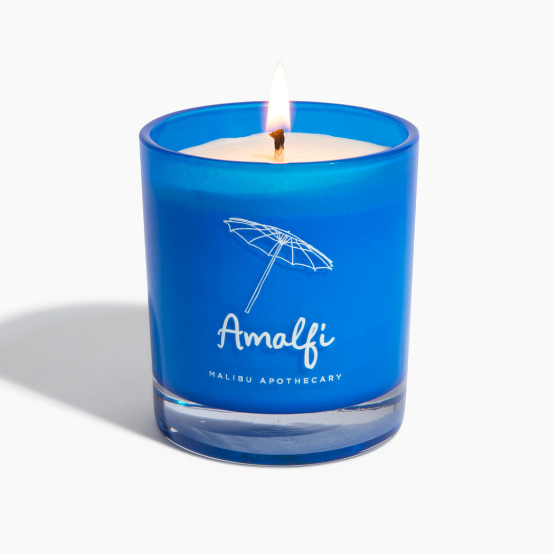 Amalfi candle burning in bright navy blue glass jar with umbrella logo against white backdrop, made with all natural coconut and soy wax and infused with basil and arugula fragrance notes