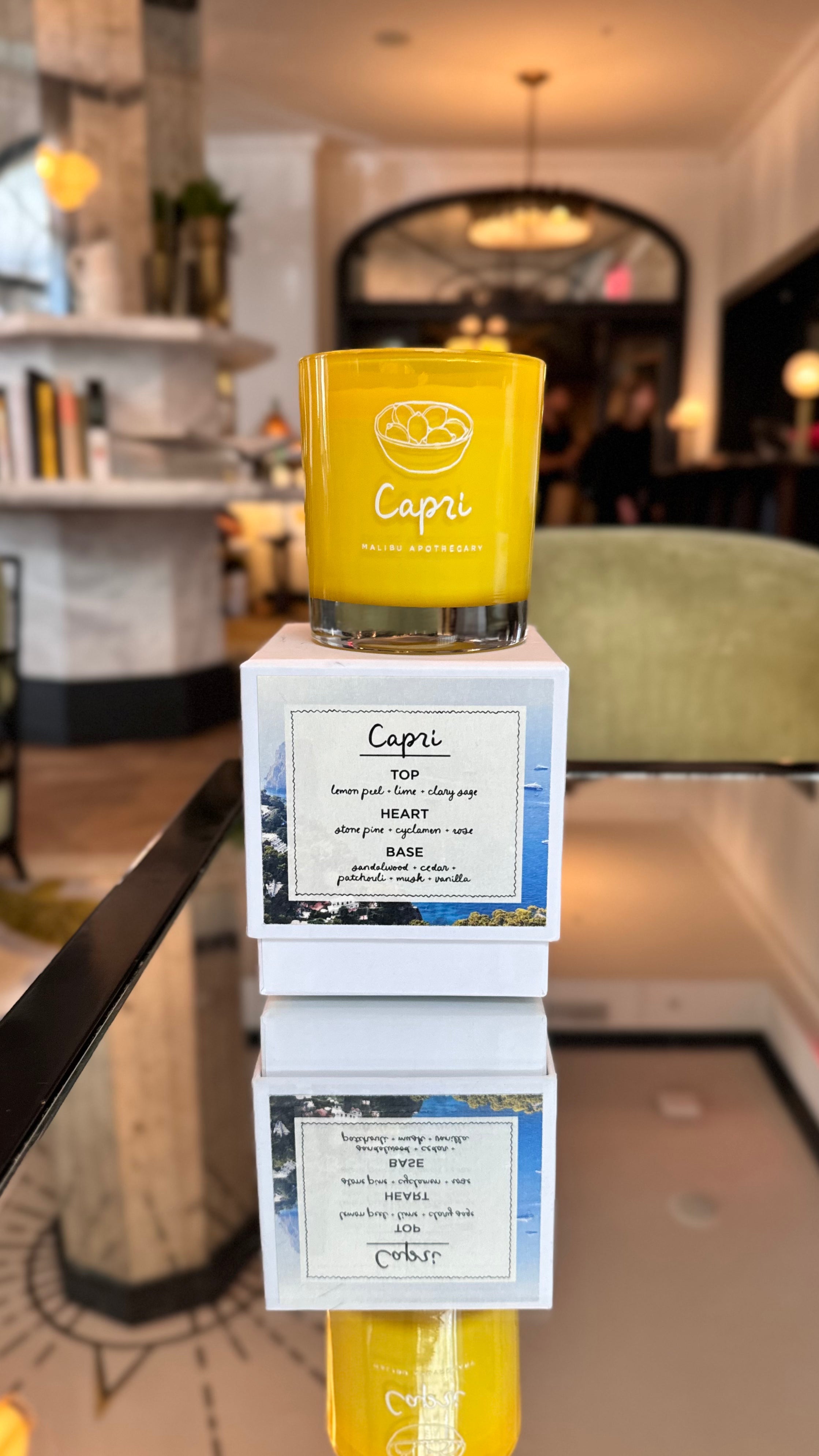 Capri candle in yellow glass jar stacked on Capri packaging boxes with fragrance note cards, displayed in a chic boutique lounge with upholstered chairs and arches; lemon peel and sage scent made with all natural coconut and soy wax