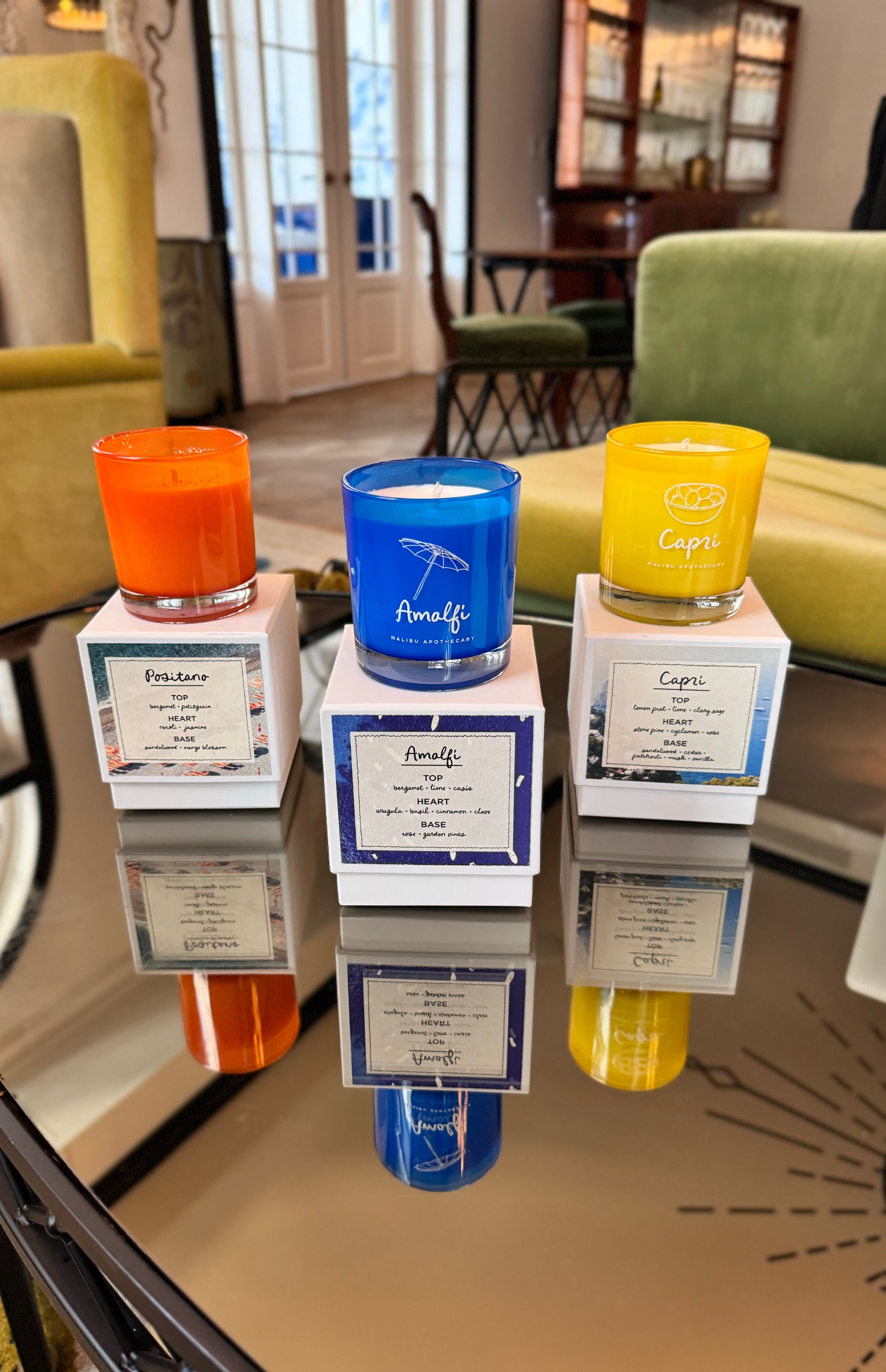 Trio of Malibu Apothecary candles—orange Positano, blue Amalfi and yellow Capri—each resting atop their scent boxes on a reflective glass table in a stylish boutique lounge; all made with all natural coconut and soy wax and clean fragrance oils