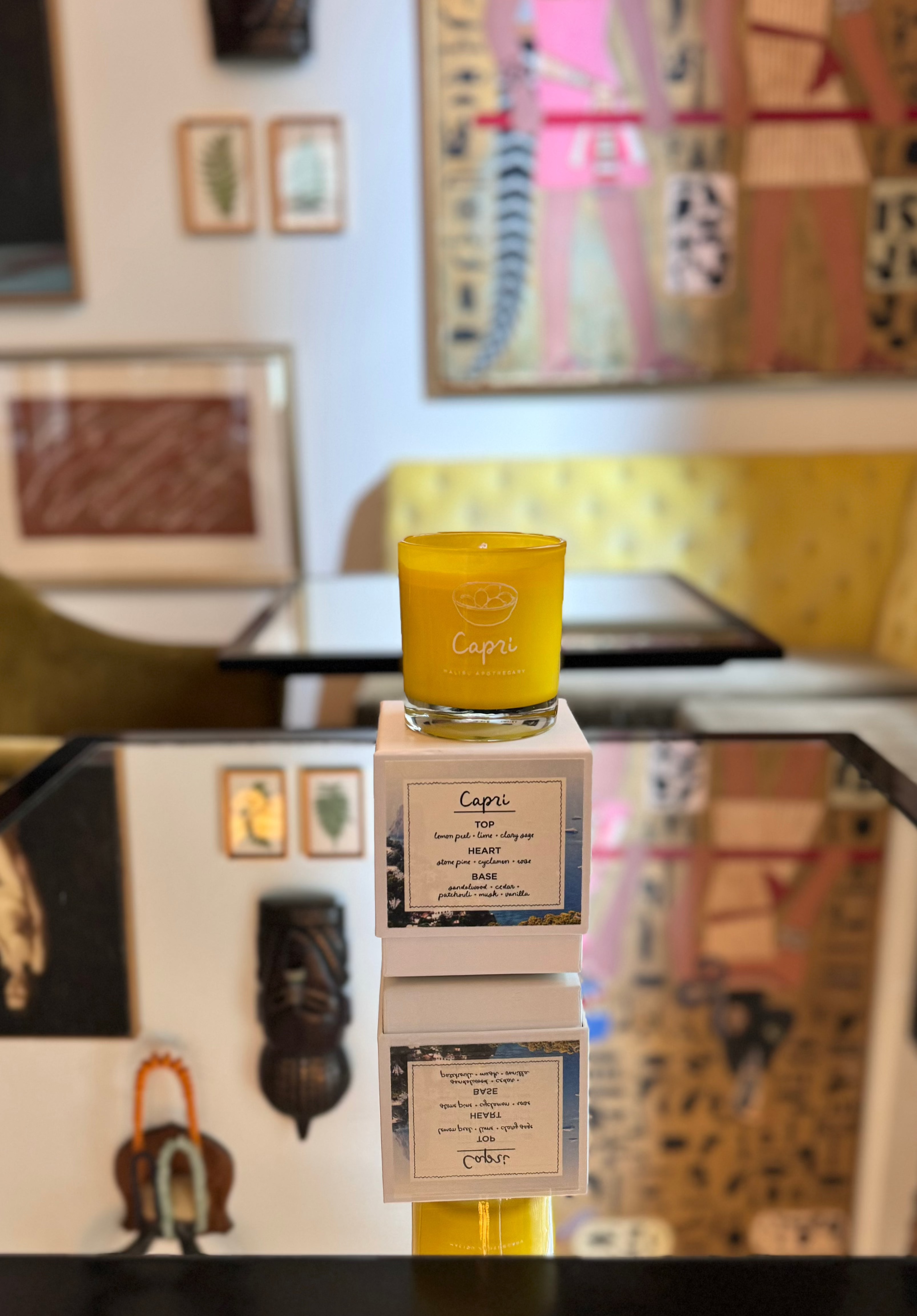 Capri candle in bright yellow glass jar stacked on matching boxes inside an eclectic art-filled room, featuring notes of sage, lemon peel and cyclamen, made with all natural coconut and soy wax