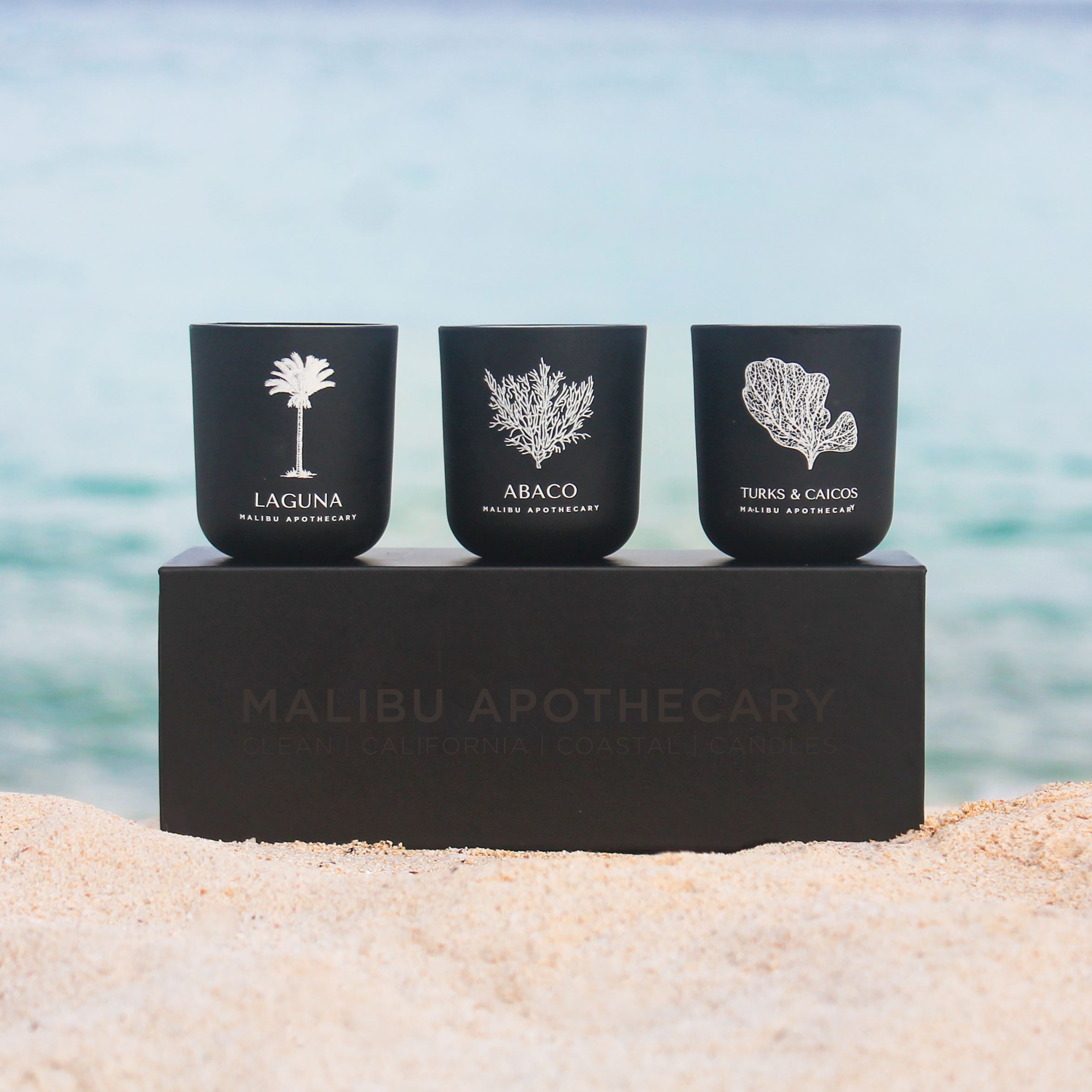 Luxury votive discovery set featuring three matte black candles hand-poured in Dallas with all natural coconut and soy wax, displayed on a sandy beach with turquoise ocean backdrop for clean fragrance inspiration