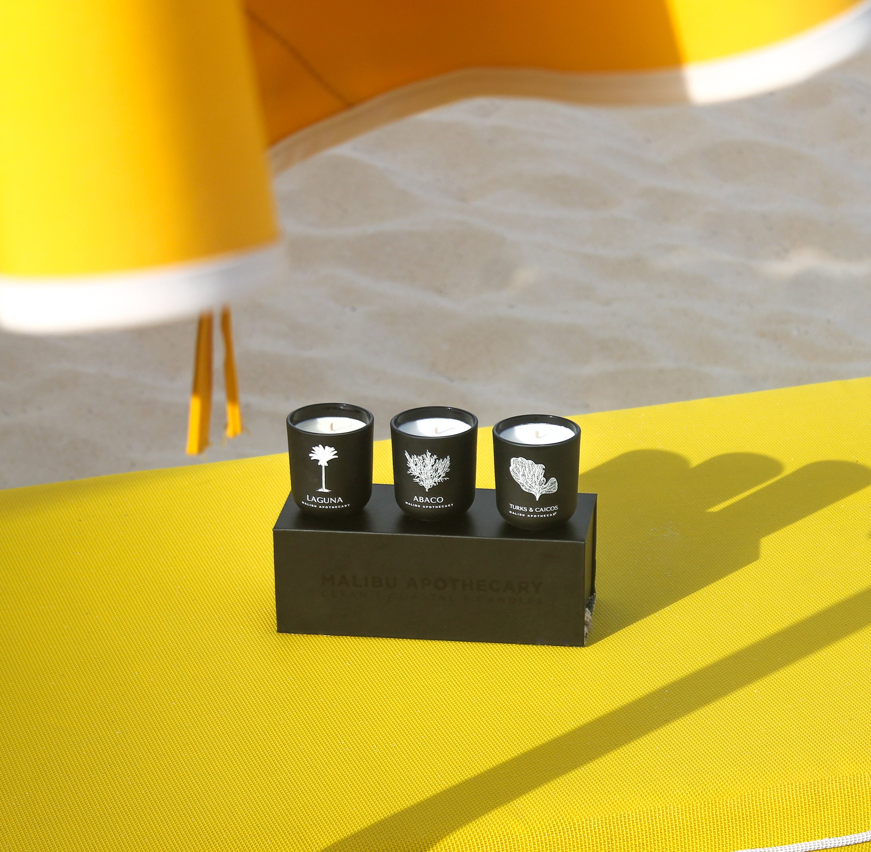 Luxury votive discovery set with three matte black candles on a black box placed on bright yellow beach towel, hand-poured with all natural coconut and soy wax for outdoor luxury fragrance