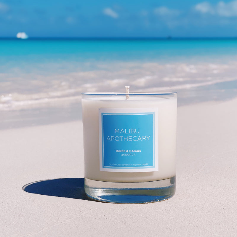 Malibu Apothecary Candle in Clear Gloss and Blue label on the sand in front of the waves in Turks & Caicos
